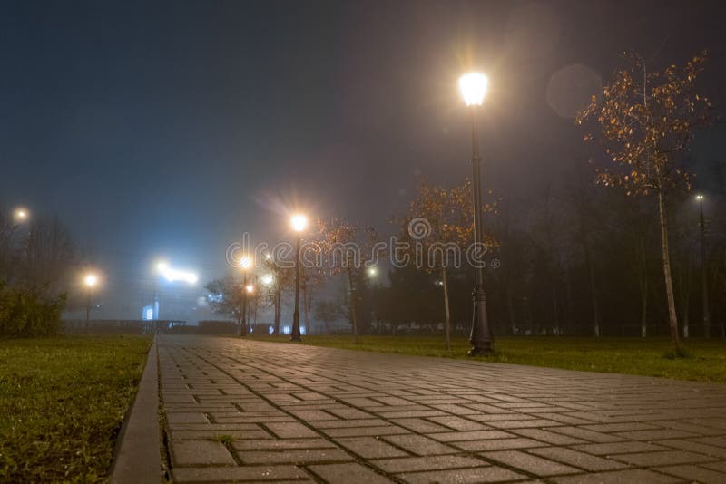 Voetpad in stadspark bij nacht in mist met straatlantaarns Mooie mistige avond in de de herfststeeg met het branden van lantaarns royalty-vrije stock foto's Voetpad in stadspark bij nacht in mist met straatlantaarns Mooie mistige avond in de de herfststeeg met het branden van lantaarns royalty-vrije stock foto's