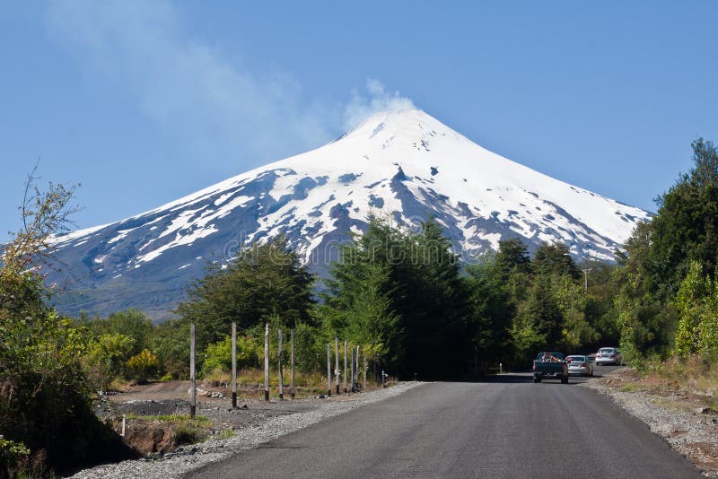 The active volcano Villarrica smoking with its snow peak framed by pine trees, and a small road with three cars. Pucon, lake region, south of Chile. The active volcano Villarrica smoking with its snow peak framed by pine trees, and a small road with three cars. Pucon, lake region, south of Chile.