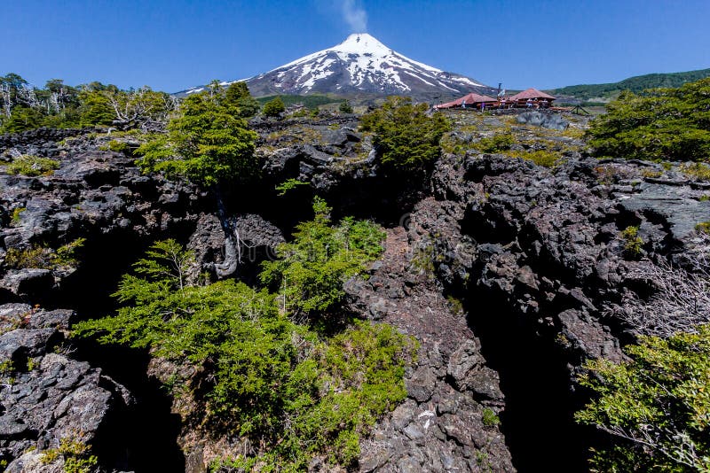 The active volcano Villarrica smoking with its snow peak. At the first plane, a lava cave with trees. Pucon, lake region, south of Chile. The active volcano Villarrica smoking with its snow peak. At the first plane, a lava cave with trees. Pucon, lake region, south of Chile.