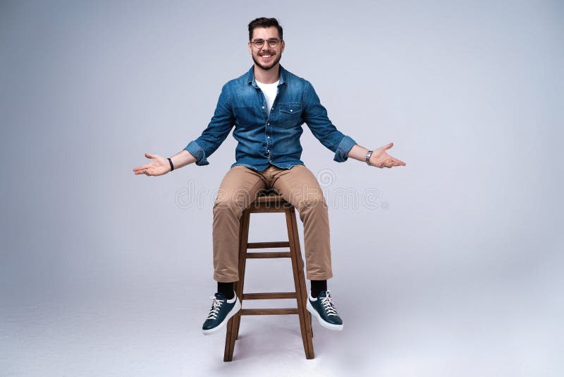 Volledig lengteportret van een aantrekkelijke jonge mens in de zitting van het jeansoverhemd op de stoel over grijze achtergrond