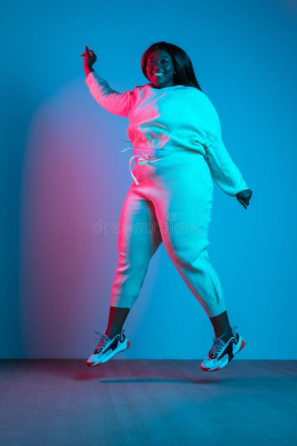 Full-length portrait of African beautiful woman with long straight hair jumping isolated on blue background in neon light. Concept of human emotions, facial expression. Bodypositive and diversity. Full-length portrait of African beautiful woman with long straight hair jumping isolated on blue background in neon light. Concept of human emotions, facial expression. Bodypositive and diversity.
