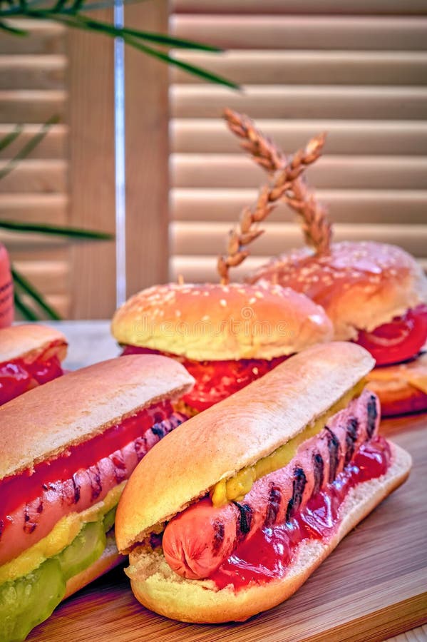 Vorbereiten von selbst gemachten Hotdogen mit Senf, Ketschup, Essiggurken und gebratenen Zwiebeln auf Holztisch Hamburger im Hint lizenzfreie stockbilder Vorbereiten von selbst gemachten Hotdogen mit Senf, Ketschup, Essiggurken und gebratenen Zwiebeln auf Holztisch Hamburger im Hint lizenzfreie stockbilder