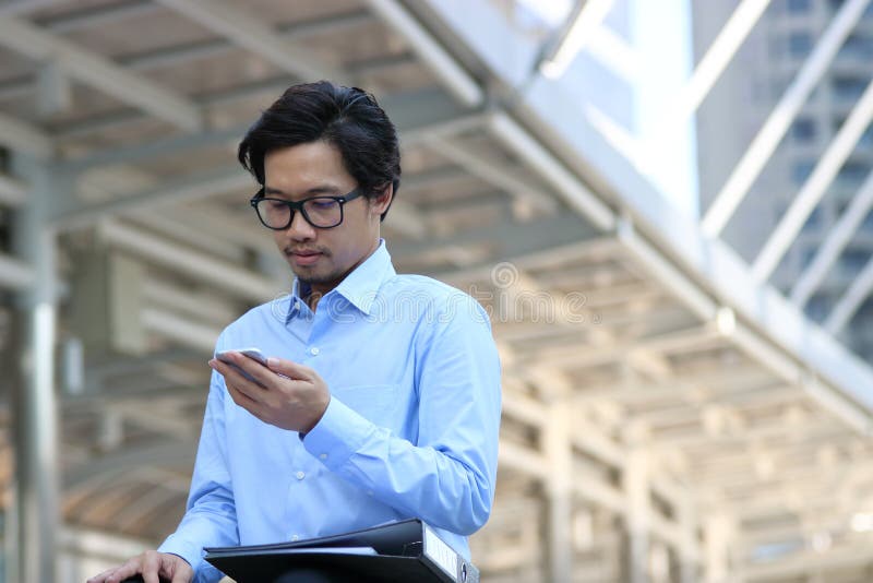Vorderansicht des hübschen jungen asiatischen Geschäftsmannes im blauen Hemd, das intelligentes Mobiltelefon äußerem Büro betrach stockfotos Vorderansicht des hübschen jungen asiatischen Geschäftsmannes im blauen Hemd, das intelligentes Mobiltelefon äußerem Büro betrach stockfotos