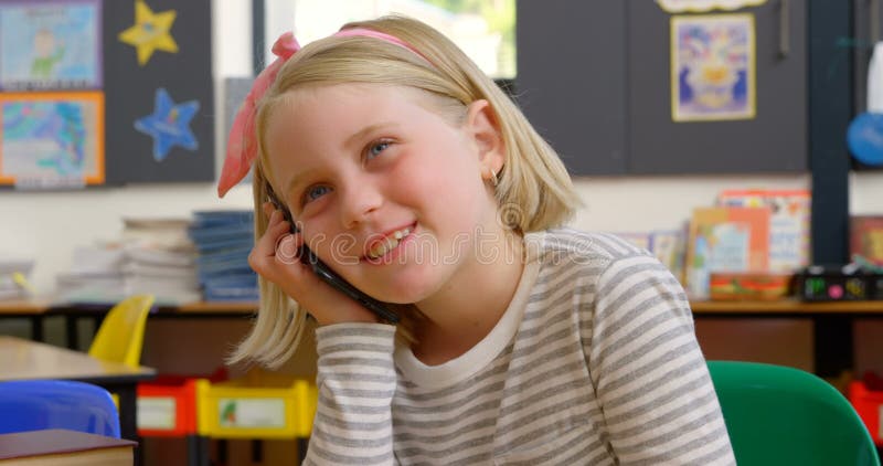 Vorderansicht des kaukasischen Schulmädchens sprechend am Handy am Schreibtisch im Klassenzimmer 4k