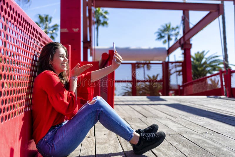 Vorderansicht einer lächelnden schönen jungen Frau, welche die städtische Kleidung sitzt auf dem Boden beim Nehmen eines selfie u lizenzfreies stockfoto Vorderansicht einer lächelnden schönen jungen Frau, welche die städtische Kleidung sitzt auf dem Boden beim Nehmen eines selfie u lizenzfreies stockfoto