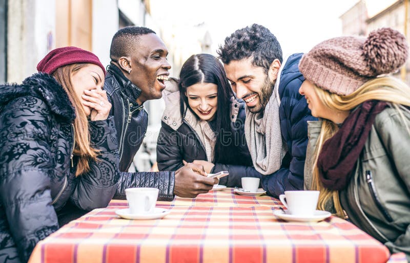 Multi-ethnic group of friends sitting in a bar and drinking coffee and watching a funny video on a cellular phone - Cheerful students meeting in a coffee house for a break. Multi-ethnic group of friends sitting in a bar and drinking coffee and watching a funny video on a cellular phone - Cheerful students meeting in a coffee house for a break