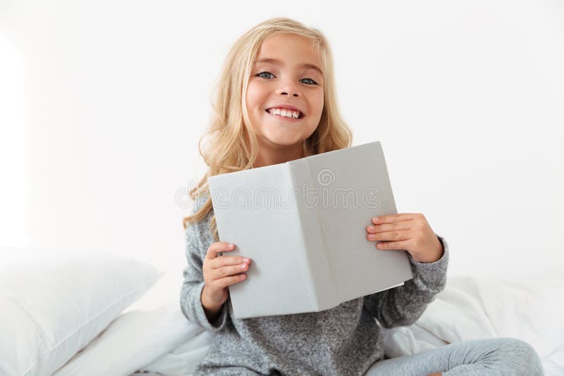 Vrolijk meisje die in grijze pyjama's boek houden, die ca bekijken royalty-vrije stock afbeeldingen Vrolijk meisje die in grijze pyjama's boek houden, die ca bekijken royalty-vrije stock afbeeldingen