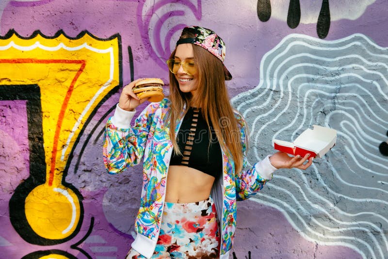 Vrolijk meisje een cheeseburger houden die zich bevindt dichtbij graffitimuur royalty-vrije stock foto Vrolijk meisje een cheeseburger houden die zich bevindt dichtbij graffitimuur royalty-vrije stock foto