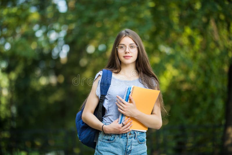 Vrolijke aantrekkelijke jonge vrouw met rugzak en notitieboekjes die en zich in park bevinden glimlachen stock foto Vrolijke aantrekkelijke jonge vrouw met rugzak en notitieboekjes die en zich in park bevinden glimlachen stock foto