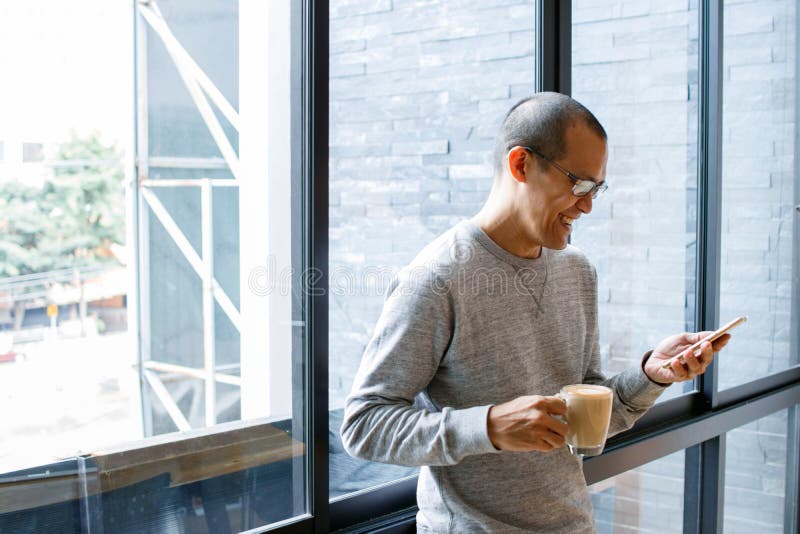 Vrolijke Aziatische mannelijke kleine bedrijfseigenaar met koffiekop in handen die op telefoon zoeken, die inkomend smsbericht op stock afbeelding Vrolijke Aziatische mannelijke kleine bedrijfseigenaar met koffiekop in handen die op telefoon zoeken, die inkomend smsbericht op stock afbeelding