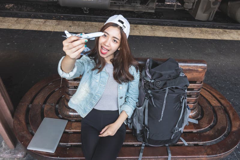 Vrolijke jonge Aziatische vrouwenreiziger met modeltreinzitting die op bank op het aankomen trein bij post wachten stock foto's Vrolijke jonge Aziatische vrouwenreiziger met modeltreinzitting die op bank op het aankomen trein bij post wachten stock foto's