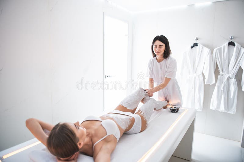 Vrolijke jonge masseur die massage met witte klei op vrouwenbenen doen royalty-vrije stock foto Vrolijke jonge masseur die massage met witte klei op vrouwenbenen doen royalty-vrije stock foto