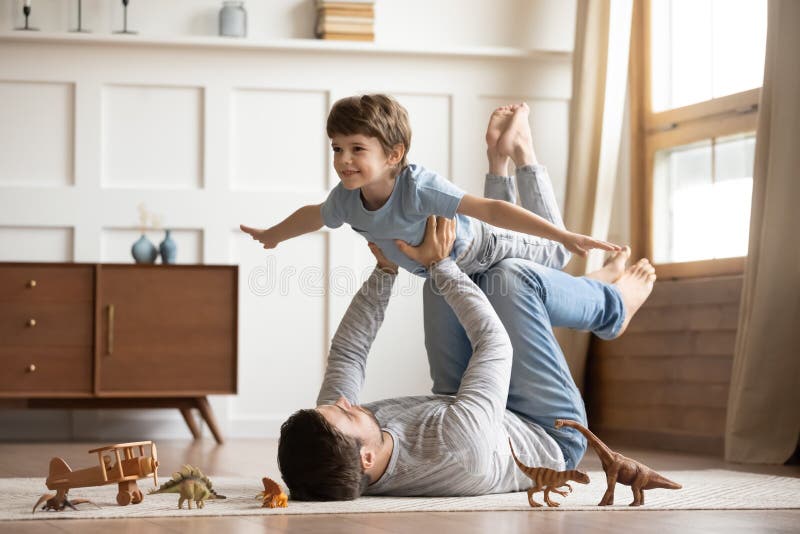 Joyful young men father lying on carpet floor, lifting excited happy little child son at home. Full length carefree two generations family having fun, practicing acroyoga in pair in living room. Joyful young men father lying on carpet floor, lifting excited happy little child son at home. Full length carefree two generations family having fun, practicing acroyoga in pair in living room.