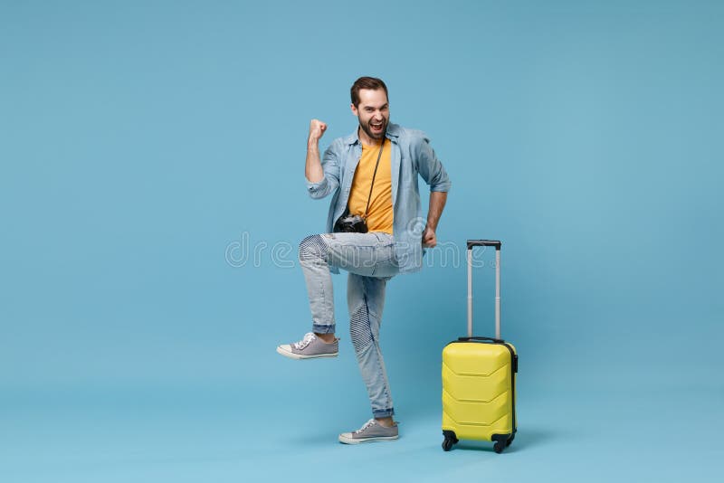 Joyful traveler tourist man in yellow clothes with photo camera, suitcase isolated on blue background. Male passenger traveling abroad on weekends. Air flight journey concept. Doing winner gesture. Joyful traveler tourist man in yellow clothes with photo camera, suitcase isolated on blue background. Male passenger traveling abroad on weekends. Air flight journey concept. Doing winner gesture