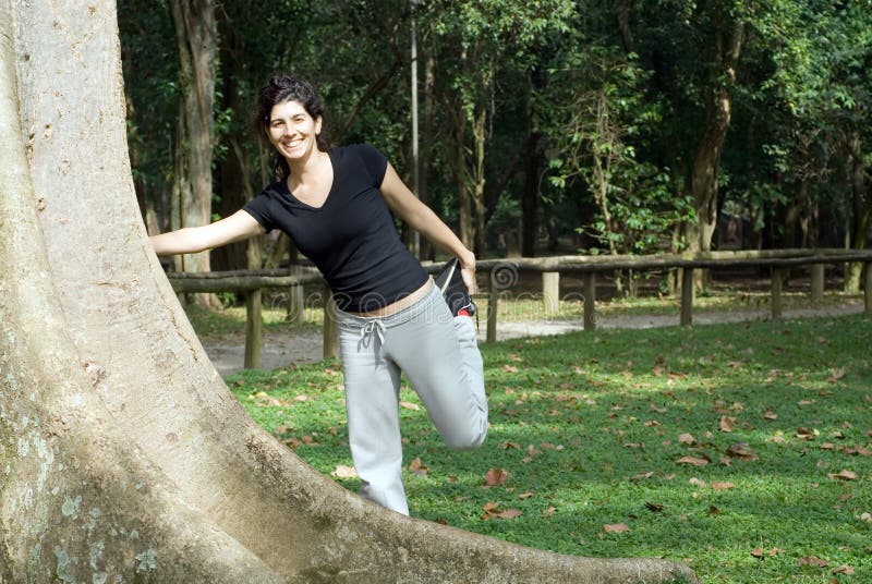 A young, attractive woman is standing next to a tree in the park. She is smiling and stretching her hamstring. Horizontally framed photo. A young, attractive woman is standing next to a tree in the park. She is smiling and stretching her hamstring. Horizontally framed photo.