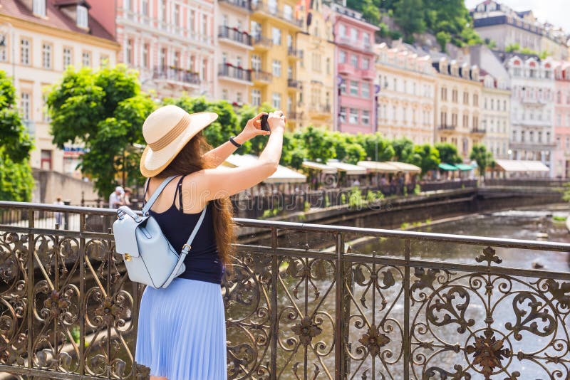 Woman taking pictures with smartphone. Stylish summer traveler woman in hat with camera outdoors in european city, old town Karlovy Vary in the background, Czech Republic, Europe. Woman taking pictures with smartphone. Stylish summer traveler woman in hat with camera outdoors in european city, old town Karlovy Vary in the background, Czech Republic, Europe.