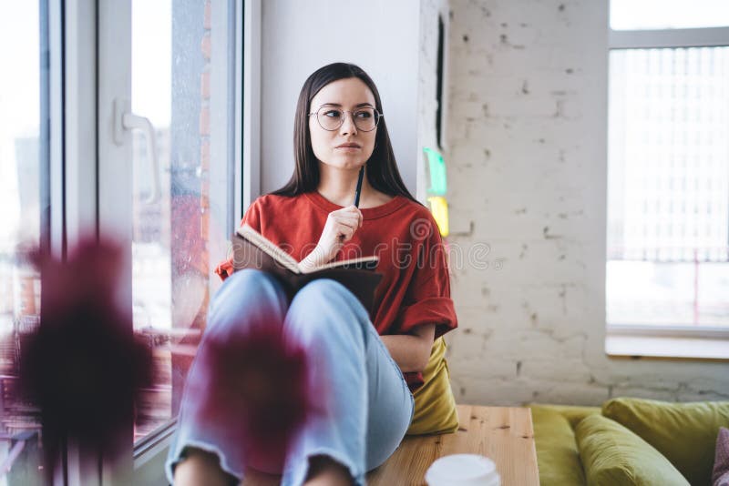 Mindful female in glasses wearing casual clothes writing notes in diary while sitting on windowsill next to flowers in modern apartment. Mindful female in glasses wearing casual clothes writing notes in diary while sitting on windowsill next to flowers in modern apartment