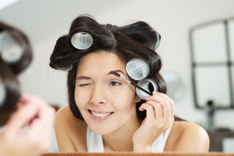 Woman in hair curlers applying eye makeup in the bathroom mirror contouring her eyebrows with eyeliner. Woman in hair curlers applying eye makeup in the bathroom mirror contouring her eyebrows with eyeliner