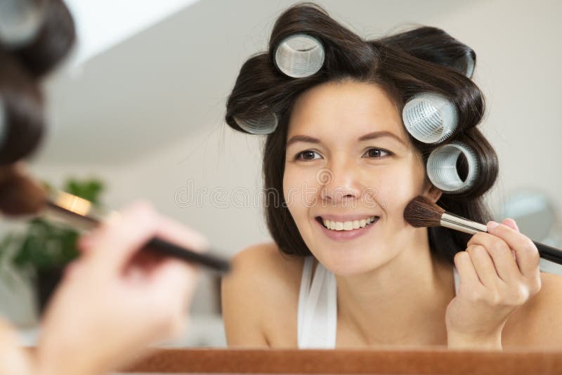 Attractive smiling woman with her long brunette hair done up in large curlers applying makeup in a mirror leaning forwards to see better as she applies blusher with a large soft cosmetics brush. Attractive smiling woman with her long brunette hair done up in large curlers applying makeup in a mirror leaning forwards to see better as she applies blusher with a large soft cosmetics brush.