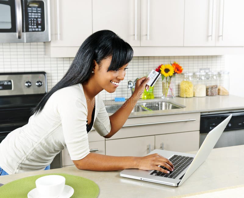 Smiling black woman online shopping using computer and credit card in kitchen. Smiling black woman online shopping using computer and credit card in kitchen