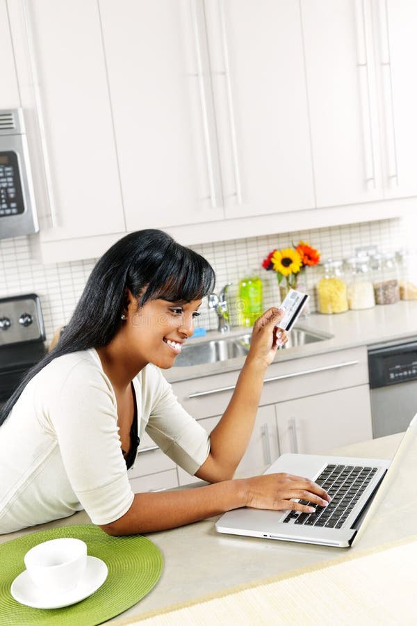 Smiling black woman online shopping using computer and credit card in kitchen. Smiling black woman online shopping using computer and credit card in kitchen