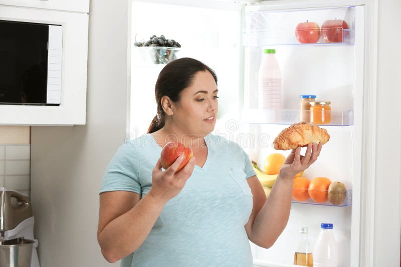 Vrouw die tussen appel en croissant dichtbij koelkast bij keuken kiezen stock afbeelding Vrouw die tussen appel en croissant dichtbij koelkast bij keuken kiezen stock afbeelding