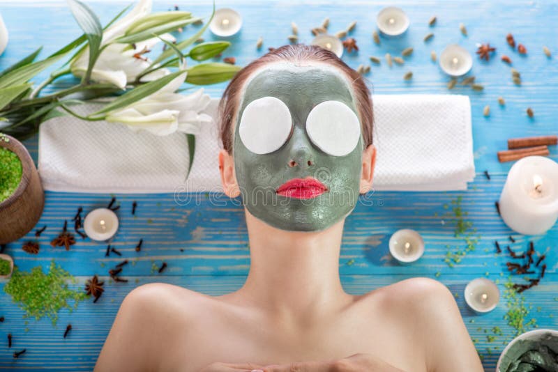 Young woman with spa facial mask on her face and cotton tampons on her eyes lying on blue table with flower, candles and sea salt in the beaty salon. Young woman with spa facial mask on her face and cotton tampons on her eyes lying on blue table with flower, candles and sea salt in the beaty salon