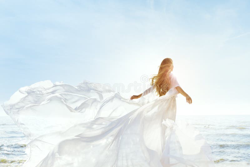 Vrouw op zonnig zeestrand in white fluttering jurk modemodel achteraan zicht zijdedoek zwaait op wind