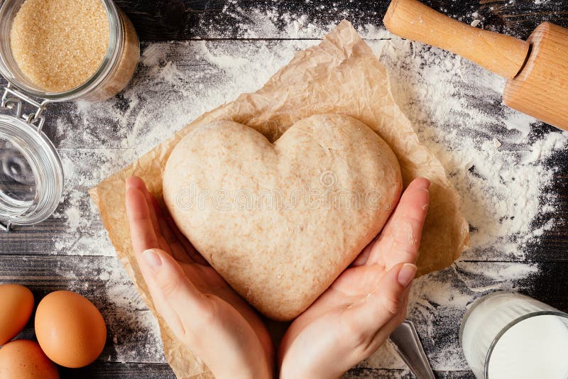 Vrouwelijke handen die deeg in de hoogste mening van de hartvorm houden Bakselingrediënten op de donkere houten lijst royalty-vrije stock afbeelding Vrouwelijke handen die deeg in de hoogste mening van de hartvorm houden Bakselingrediënten op de donkere houten lijst royalty-vrije stock afbeelding