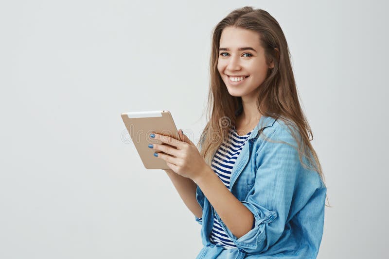 Feminine tender gorgeous caucasian young woman wearing trendy clothes smiling delighted friendly holding digital tablet, enjoying weekend messaging friends reading online book, white background. Feminine tender gorgeous caucasian young woman wearing trendy clothes smiling delighted friendly holding digital tablet, enjoying weekend messaging friends reading online book, white background.