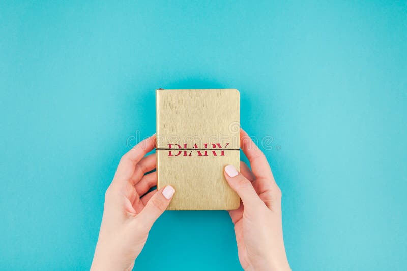 Creative flat lay of woman hands holding small golden notebook diary with copy space on turquoise paper background in minimalism style. Template for feminine blog, social media. Creative flat lay of woman hands holding small golden notebook diary with copy space on turquoise paper background in minimalism style. Template for feminine blog, social media
