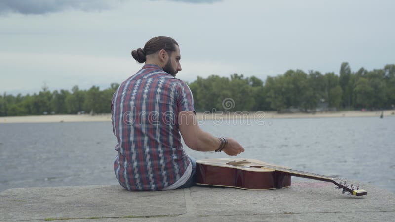Vue arrière d'homme frustrant siégeant avec la guitare sur la rive du fleuve et de lancer des pierres dans l'eau De jeunes Caucas clips vidéos