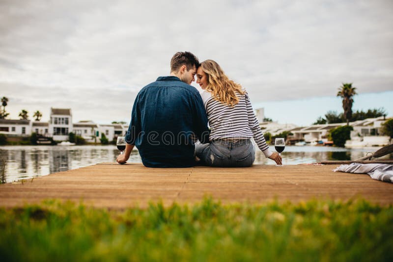 Rear view of a couple in love sitting together touching their heads near a lake. Couple on a day out sitting together holding wine glasses on the wooden dock near a lake. Rear view of a couple in love sitting together touching their heads near a lake. Couple on a day out sitting together holding wine glasses on the wooden dock near a lake.