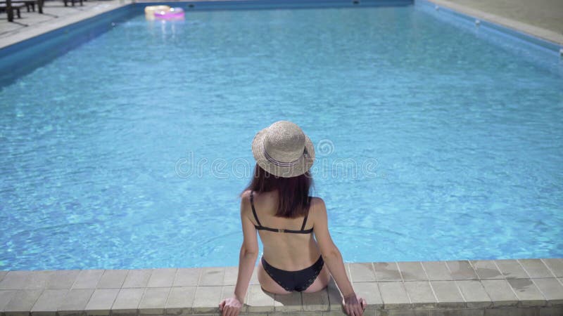 Vue arrière d'une belle fille en maillot de bain noir relaxant au bord de la piscine une femme sexy dans un bikini assise sur banque de vidéos