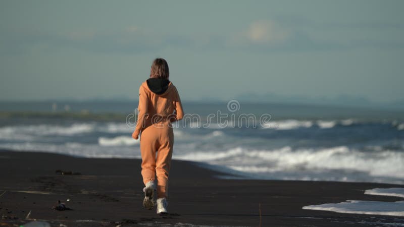 Vue arrière d'une femme en combinaison orange courant sur la plage de sable sur fond des vagues de rupture de l'océan banque de vidéos