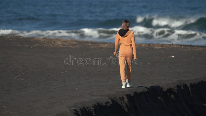Vue arrière d'une femme en combinaison orange marchant le long d'une plage de sable noir avec océan en arrière-plan banque de vidéos