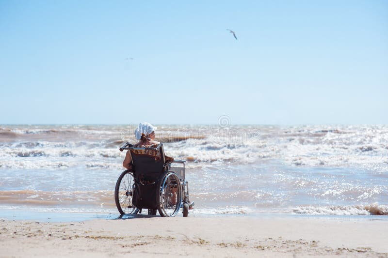 Vue arrière d'une femme handicapée s'asseyant sur un fauteuil roulant sur la plage un jour ensoleillé Elle regarde l'océan image libre de droits Vue arrière d'une femme handicapée s'asseyant sur un fauteuil roulant sur la plage un jour ensoleillé Elle regarde l'océan image libre de droits
