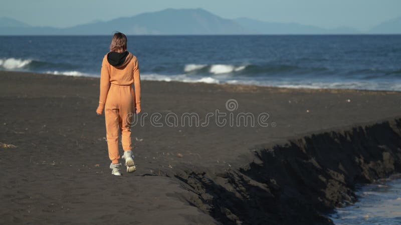 Vue arrière d'une femme méconnaissable en combinaison orange marchant sur la plage de sable noir à l'heure d'or clips vidéos