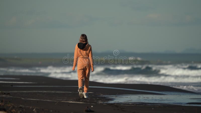 Vue arrière d'une femme méconnaissable en combinaison orange marchant sur la plage sur fond de l'océan pacifique banque de vidéos