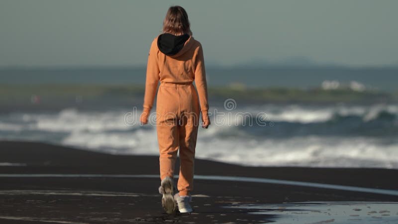 Vue arrière d'une femme méconnaissable en combinaison orange sur la plage sur fond de vagues brisantes banque de vidéos