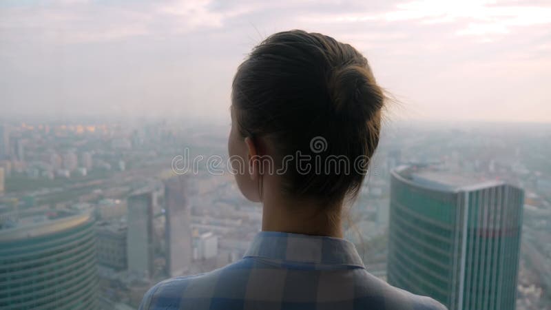 Vue arrière d'une femme pensive regardant le paysage urbain à travers la fenêtre du gratte-ciel banque de vidéos