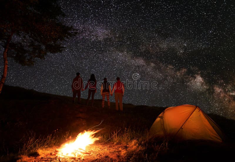 Back view of two pair hikers holding hands and enjoying milky way at starry sky during a night of camping near a campfire and orange tent in mountains. Romantic evening under the fabulous starry sky. Back view of two pair hikers holding hands and enjoying milky way at starry sky during a night of camping near a campfire and orange tent in mountains. Romantic evening under the fabulous starry sky