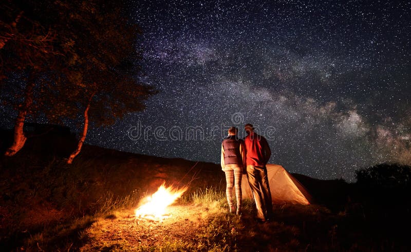 Rear view of two person backpackers clinging to each other, looking at the starry sky with milky way, near the campfire, tent and trees. Concept of active rest at night camping in the mountains. Rear view of two person backpackers clinging to each other, looking at the starry sky with milky way, near the campfire, tent and trees. Concept of active rest at night camping in the mountains