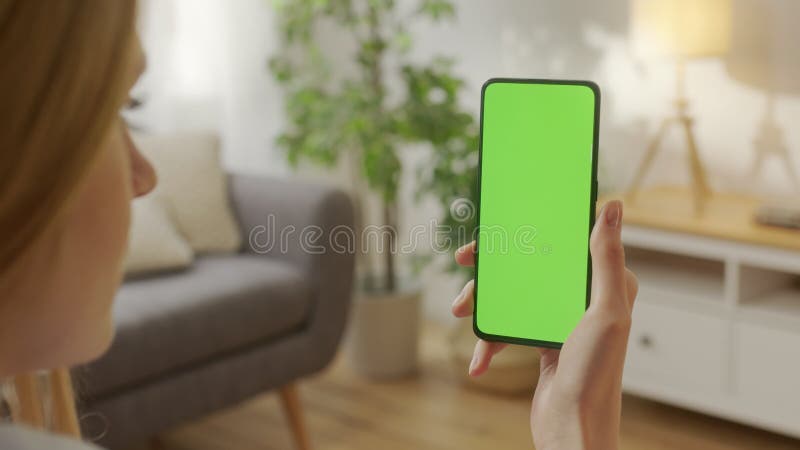 Vue arrière de la femme dans le salon en utilisant téléphone avec écran de maquette vert chroma clé sans points de piste surfer in banque de vidéos
