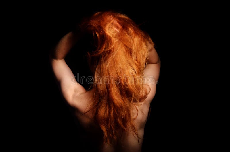 A ginger nude woman is standing back in the dark. She has raised her long red hair and turned left her face. A ginger nude woman is standing back in the dark. She has raised her long red hair and turned left her face.