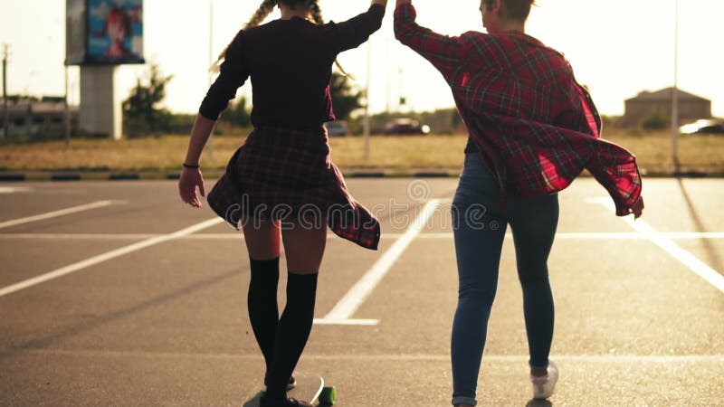 Vue arrière de la jeune fille attirante de hippie enseigné le skateboarding par un ami qui soutient sa participation sa main