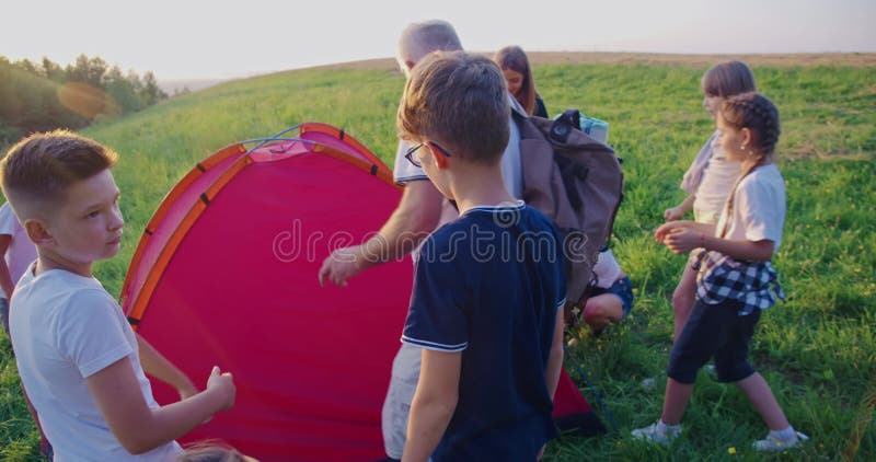 Vue arrière du groupe d'enfants monter la tente rouge organisinf campsite travailler ensemble vieux enseignant et banque de vidéos