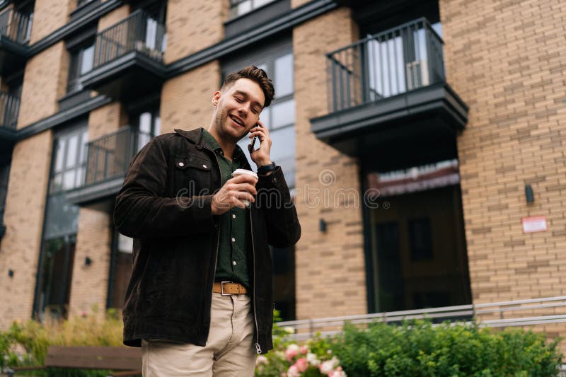 Vue basse du jeune homme élégant en vêtements décontractés tenant une tasse de café en papier et parlant sur le smartphone pendant photographie stock libre de droits