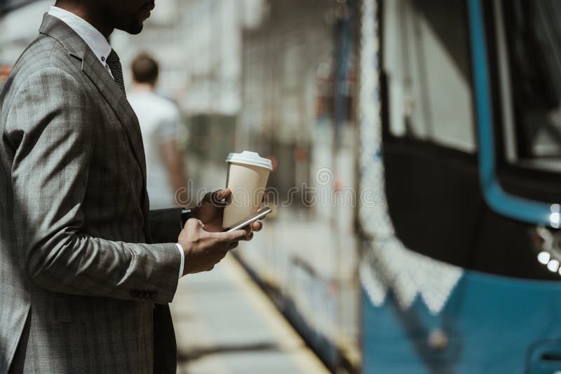 Vue cultivée de jeune homme d'affaires d'afro-américain avec la tasse de smartphone et de café prenant le train photographie stock libre de droits Vue cultivée de jeune homme d'affaires d'afro-américain avec la tasse de smartphone et de café prenant le train photographie stock libre de droits