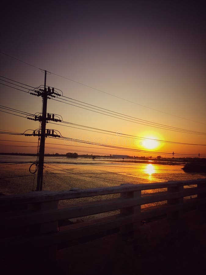 Vue de coucher du soleil de rive sur le pont et le poteau électrique dans l'eau image stock Vue de coucher du soleil de rive sur le pont et le poteau électrique dans l'eau image stock